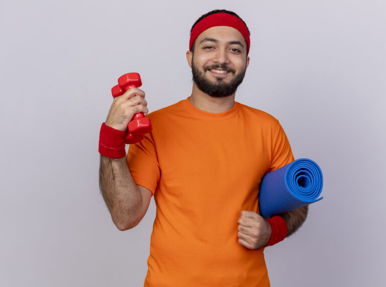 smiling young sporty man wearing headband and wristband holding dumbbells with yoga mat isolated on white background