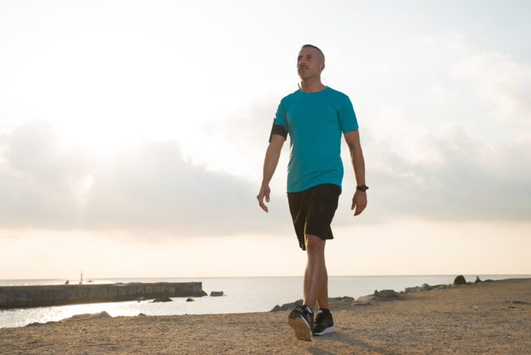 Confident male jogger walking after running on beach. Pensive young man in earphones listening to music while walking alone on shore. Recreation concept